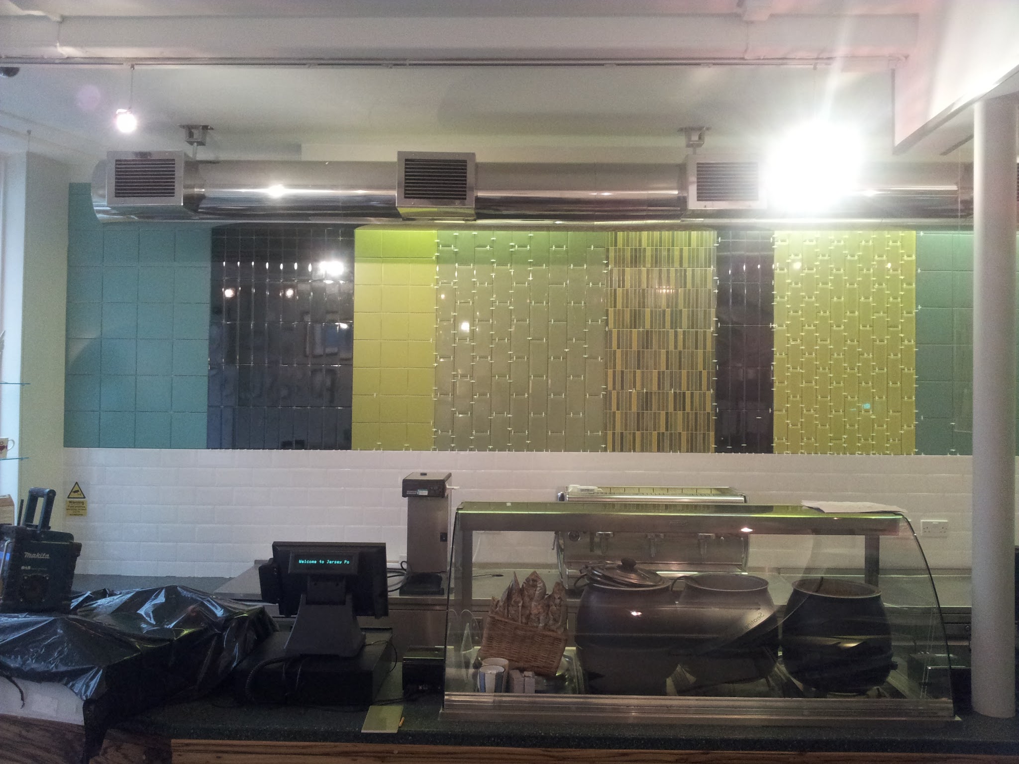 Die fertige Fliesenwand vom Servicebereich des Cafés aus gesehen. Grüne und türkise Farbtöne werden durch glänzende schwarze Flächen und ein zentrales Mosaikdetail ausgewogen ergänzt.