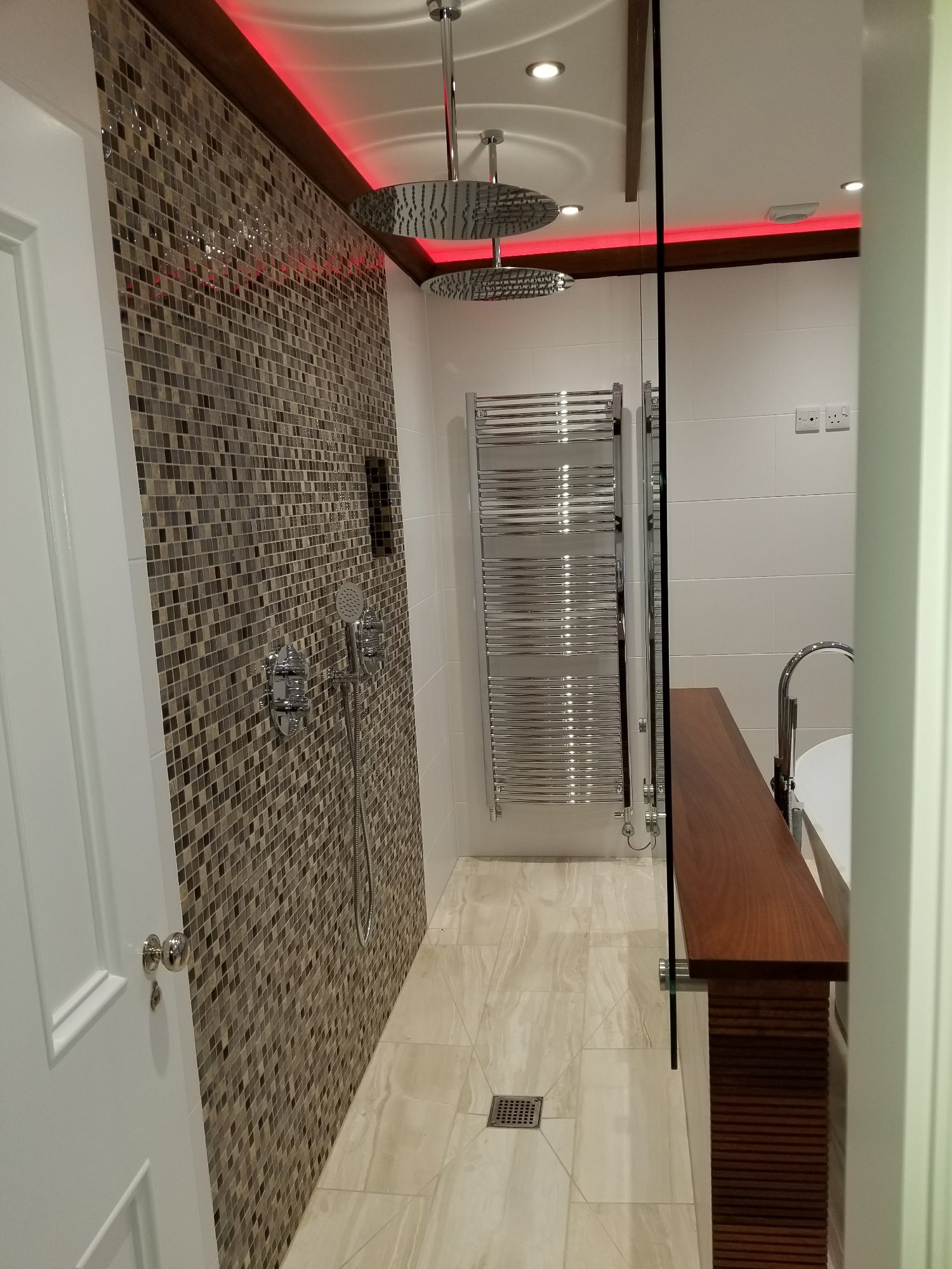 The finished shower features a mosaic wall, glass screen, dual rainfall shower heads, handheld controls and a chrome towel radiator.