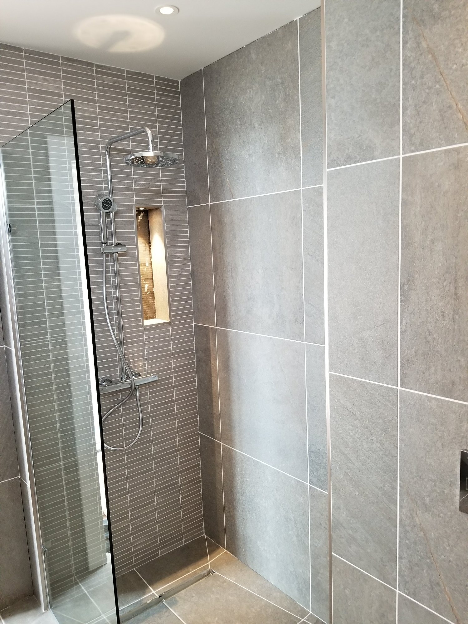 La douche terminée associe de grands carreaux gris, un mur décoratif en carreaux étroits et une paroi en verre transparent.