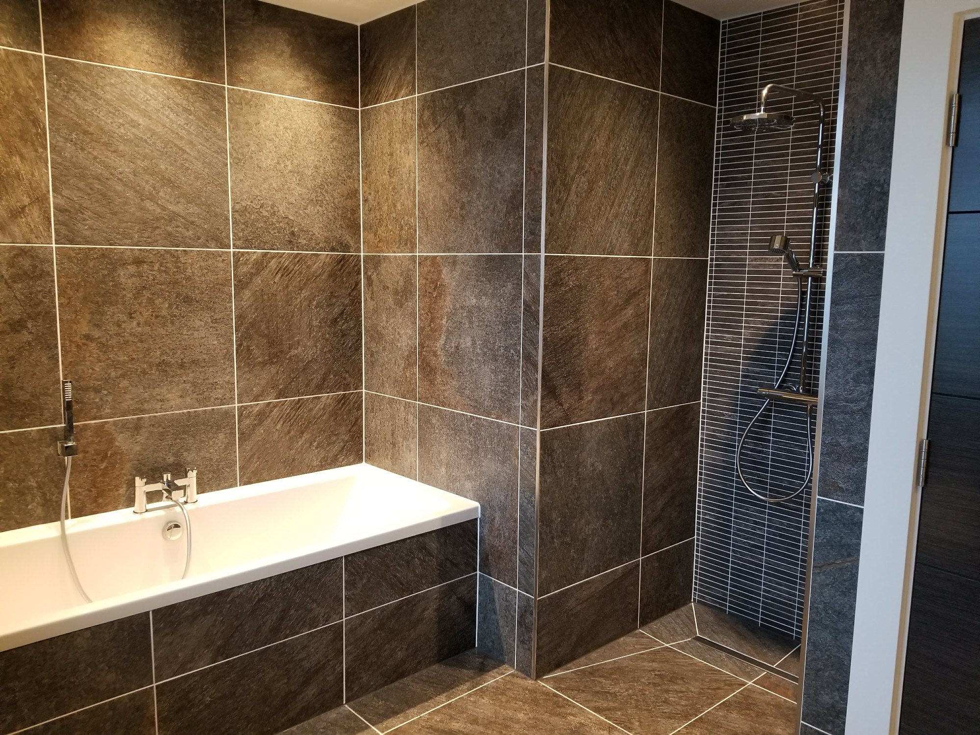 The narrow-format feature tiles highlight the rainfall shower and recessed niche while complementing the surrounding stone-effect surfaces.