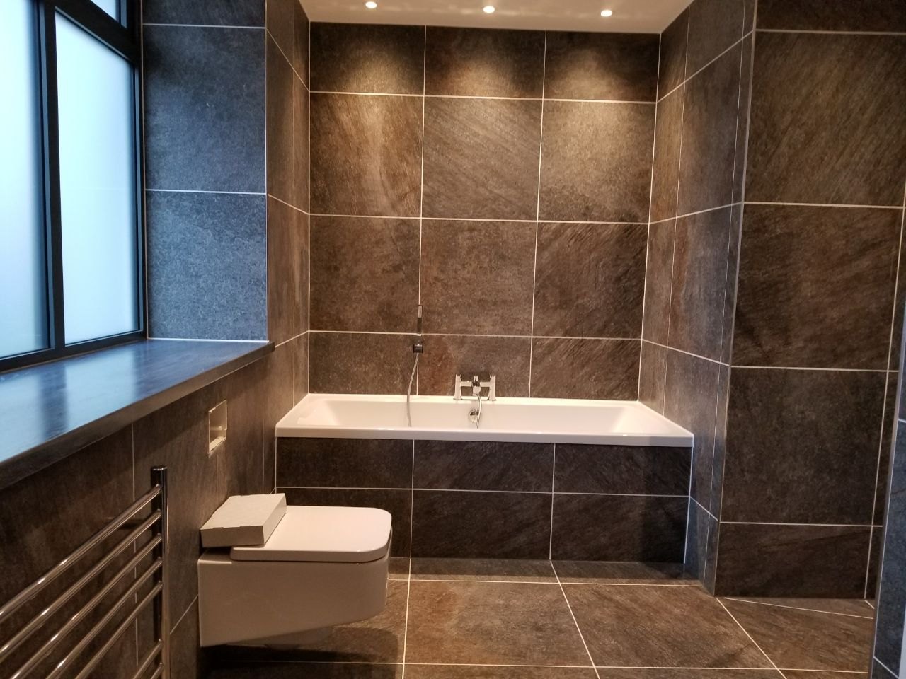 The wall-hung toilet, chrome towel radiator and fully tiled bath area create a clean and functional layout.