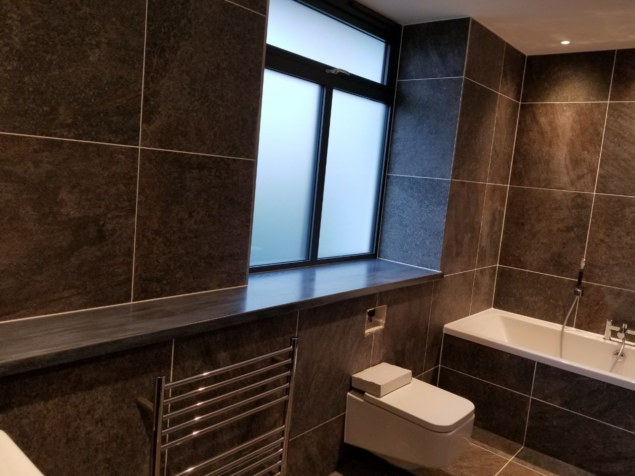 Dark stone-effect tiles, a matching window sill, wall-hung toilet and chrome towel rail give the room a consistent modern appearance.